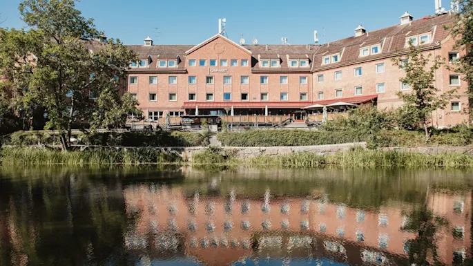 En stor mursteinsbygning med et skilt hvor det står KOMPANIET reflekteres i et rolig vann med trær og grønt løvverk.