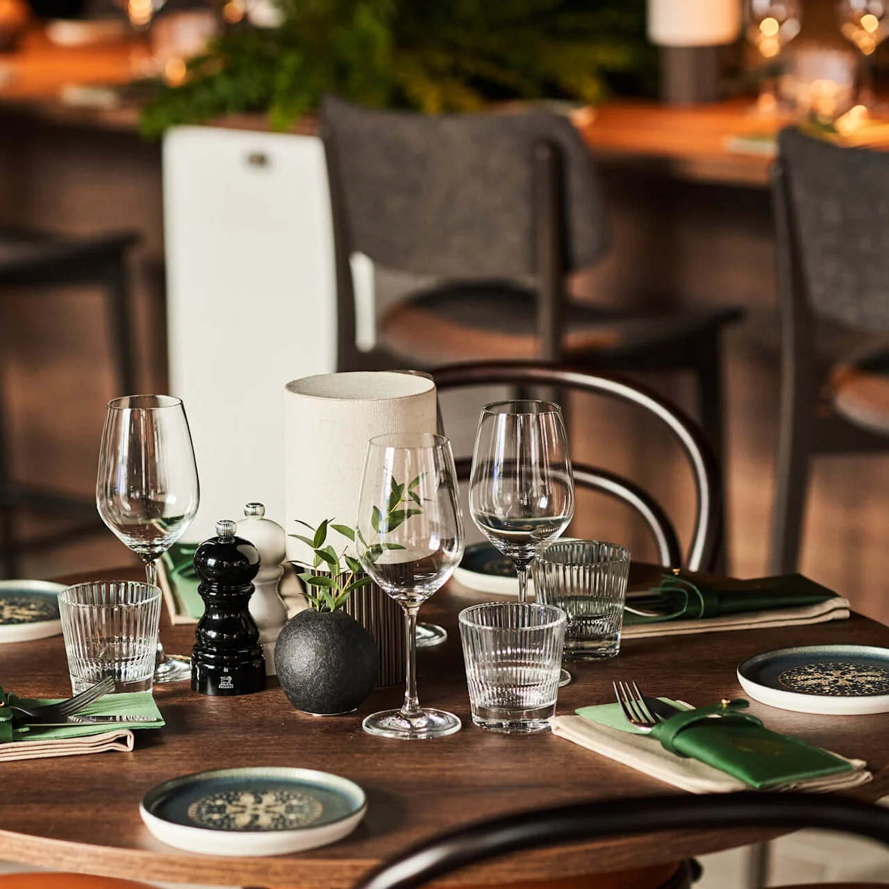 Et rundt træbord er elegant dækket med glas, tallerkener, bestik og små planter. I baggrunden ses barstole, hvilket tyder på en hyggelig restaurantstemning.