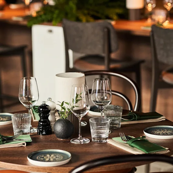 Ett runt träbord är smakfullt dukat med glas, tallrikar, bestick och små växter. I bakgrunden syns barstolar, vilket tyder på en mysig restaurangmiljö.
