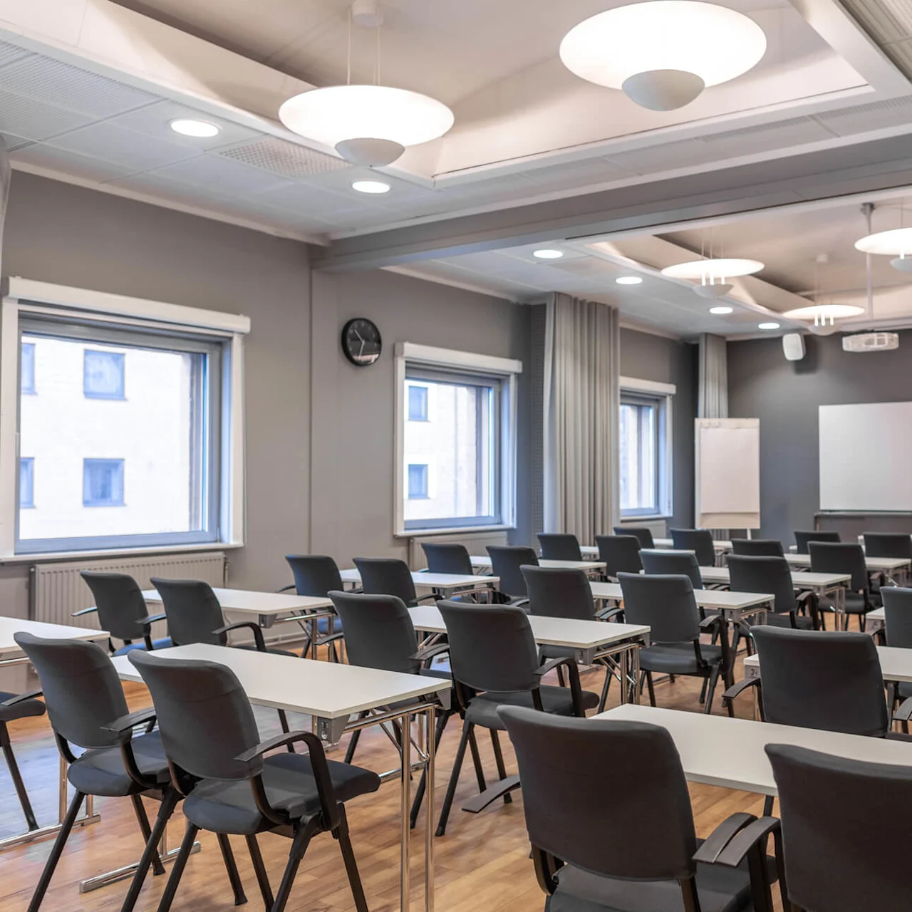 Classroom with empty desks and black chairs, arranged in rows facing a whiteboard. Windows on the left, clock on the wall, projector on the ceiling.