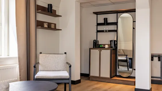 Viihtyisä hotellihuone, jossa on mukava nojatuoli, sohvapöytä ja minibaariyksikkö kokovartalopeilillä.