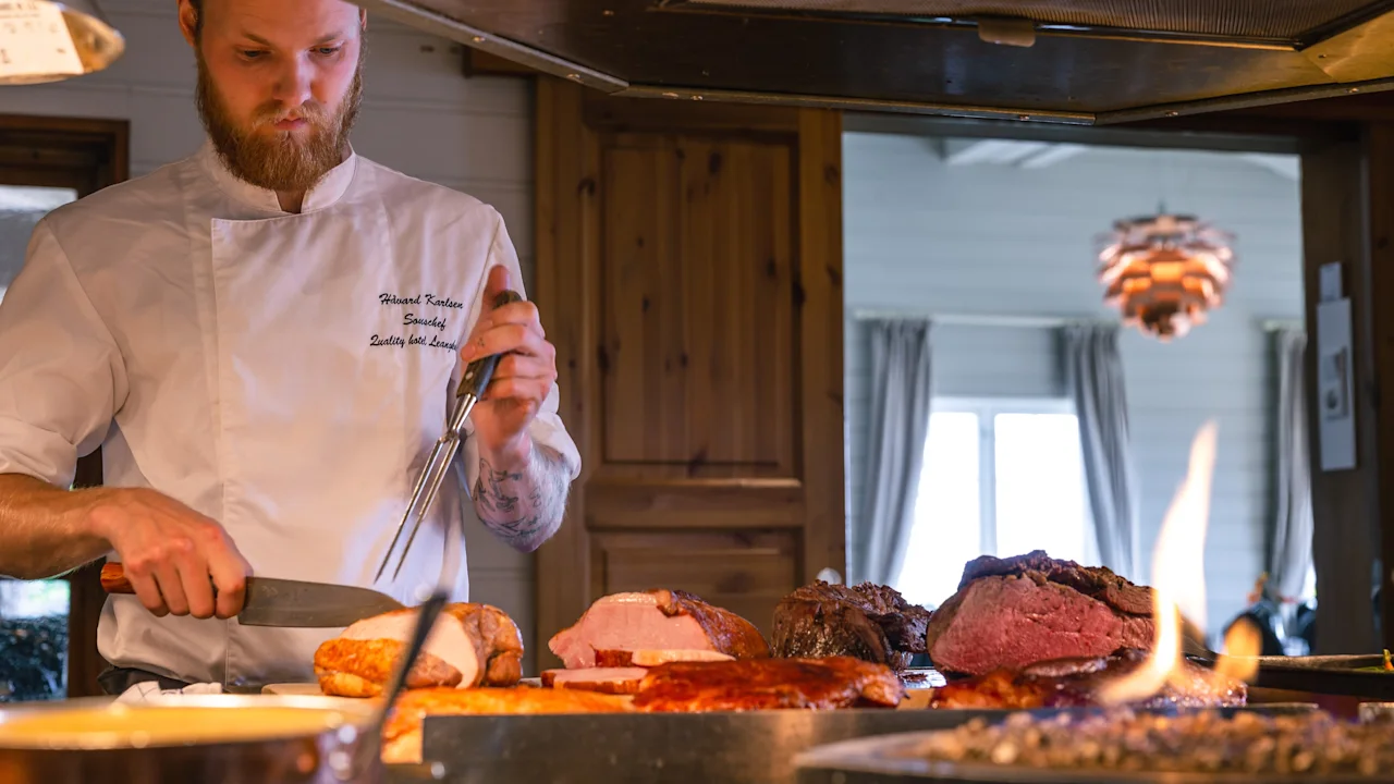 Kock skär rostade kött på disk i mysigt kök. Varm eld i förgrunden; fönster och trädörr i bakgrunden. Text på kockens jacka: "Bouche à Bouche, Quality Gastrobar."