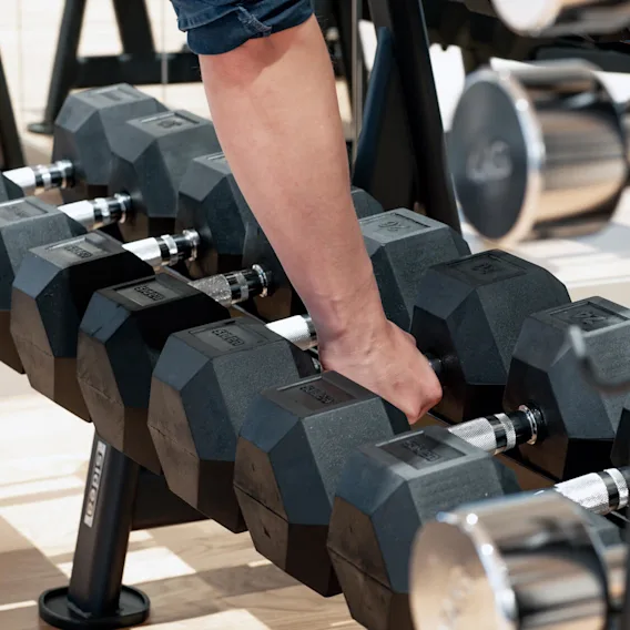 Dumbbells are being held by a person’s hand, lifting one from a rack in a gym setting. The surrounding area has additional weights and gym equipment. 