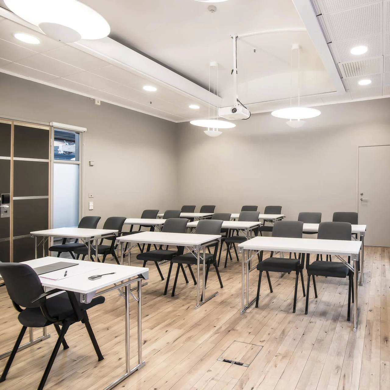 Classroom with neatly arranged tables and chairs facing a projector. Bright overhead lighting and windows illuminate the room. A side table holds supplies near a coat rack.