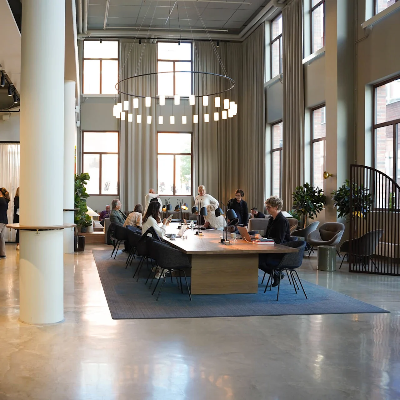 People work and socialize in a spacious, modern office cafeteria with tall windows and comfortable furnishings.