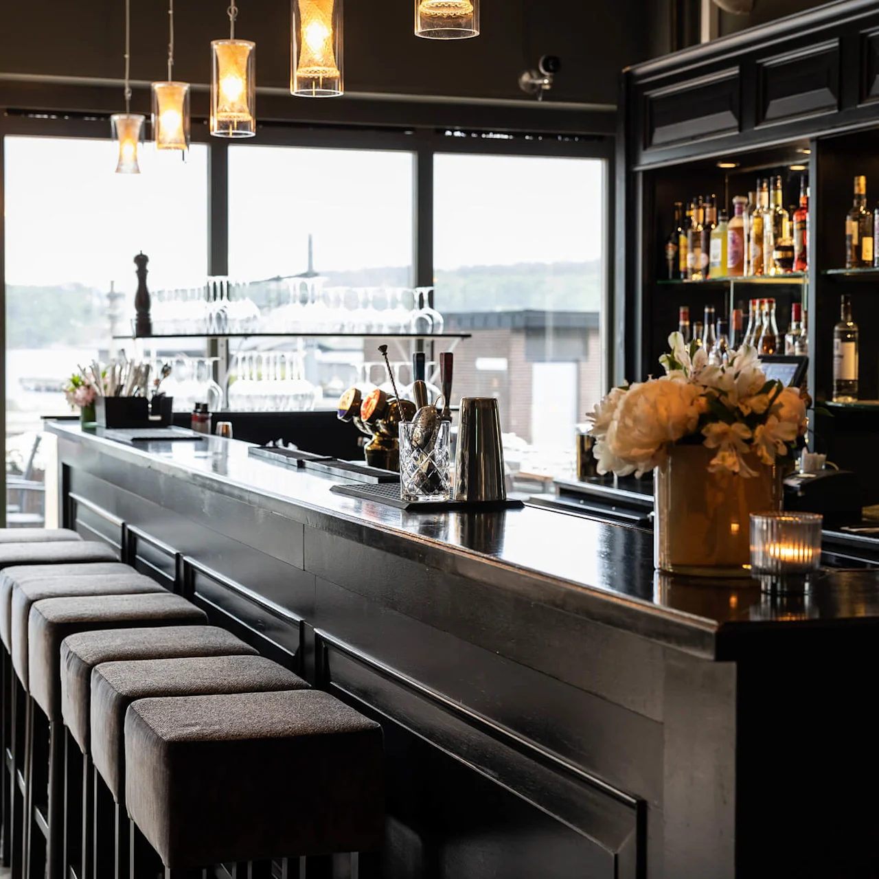 Elegant svart bardisk med høye stoler står i et svakt opplyst loungeområde. Bak er hyller med diverse flasker med brennevin. Store vinduer overser en uteplass og fjernt landskap ved vannet.