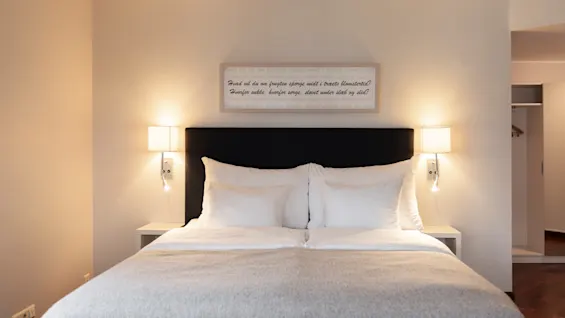 A serene hotel room featuring a comfortable bed with crisp white linens, soft lighting from wall lamps, and a framed quote above.