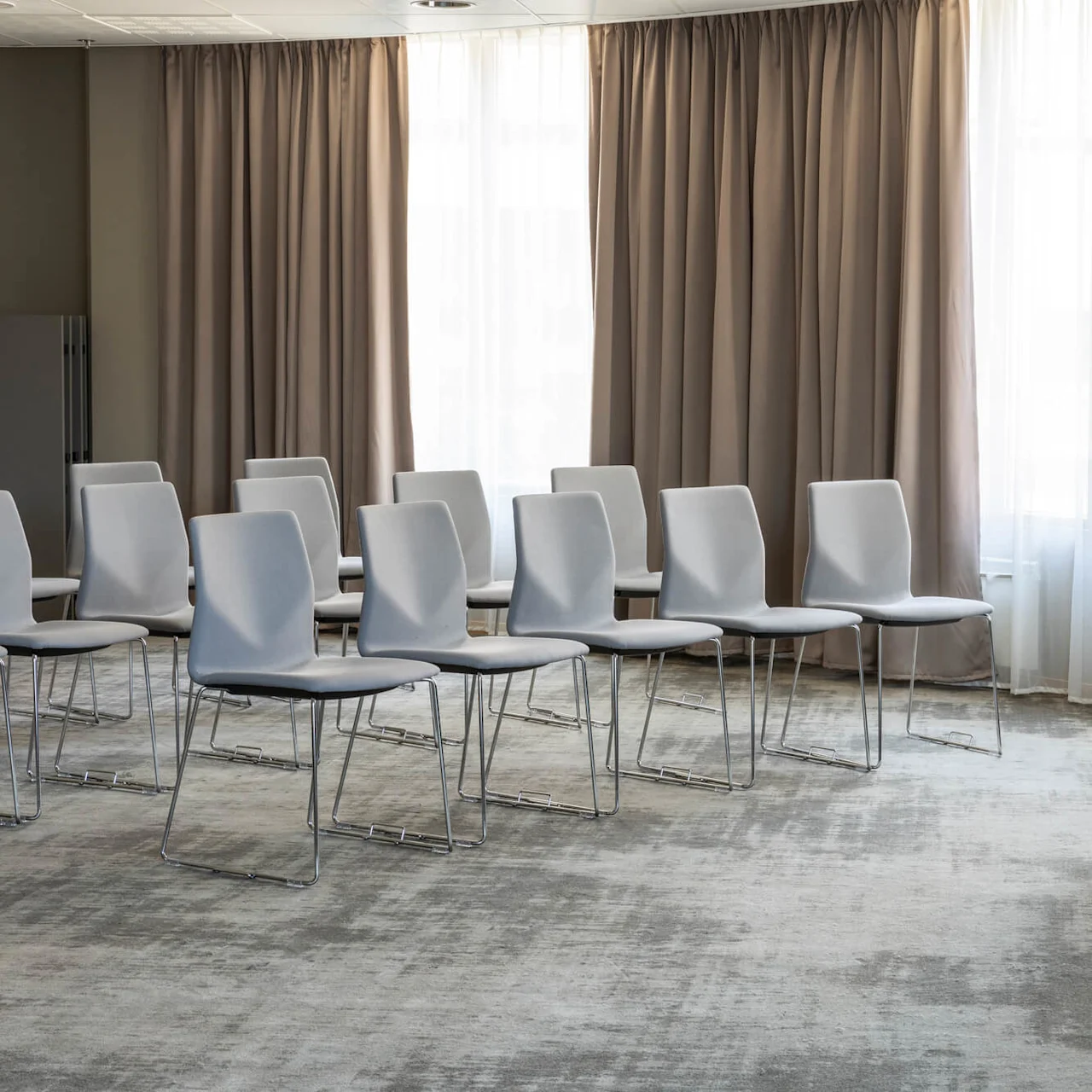 Rows of white chairs face a table with a black skirt, situated in a room with curtain-covered windows and a patterned carpet.