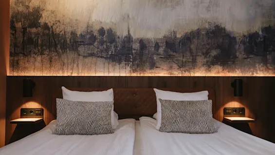 Et koselig hotellrom med en stor seng, mønstrede puter og et abstrakt maleri over sengegavlen, opplyst av varm belysning.