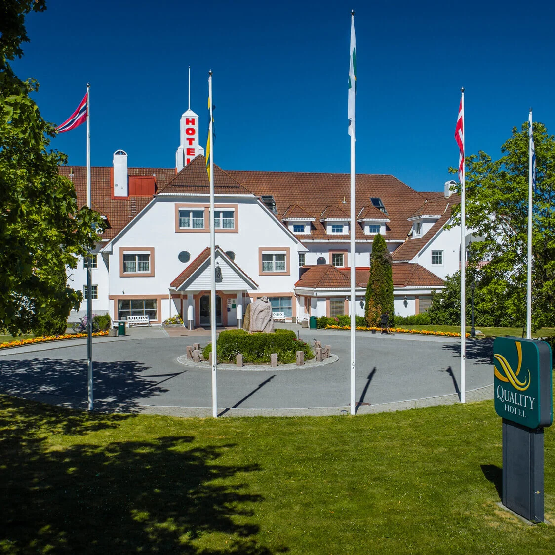 Et hvidt fler-etagers hotel med rød "HOTEL"-skilt, omgivet af træer og flag, står i solrigt vejr. En rundkørsel fører til indgangen. "QUALITY HOTEL" står på et grønt skilt.