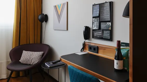 En modernt inredd arbetsyta med en stilig stol, ett skrivbord med telefon och vin, omgiven av konst och informationstavla, i en hotellrumsmiljö.