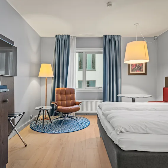 Et ryddig hotellrom med seng, tregulv, lenestol og bord under en lys taklampe. Blå gardiner dekker vinduet, og en TV er montert på veggen.