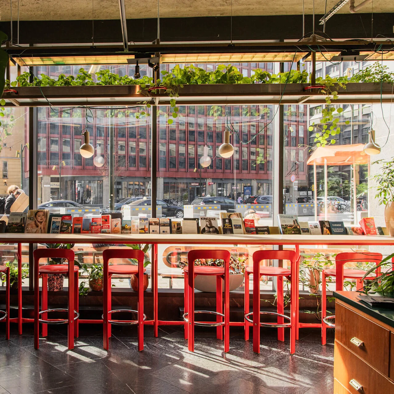 Barstools line a high table with books, framed by large windows and hanging plants. Outside, a busy city street is visible. 