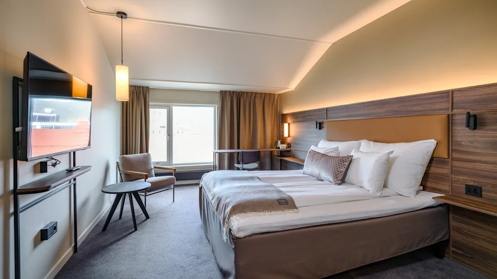 En pent gjort hotellseng med hvite laken er plassert nær et stort vindu, opplyst av myk belysning. En veggmontert TV og et lite bord med en stol fullfører det moderne rommet.