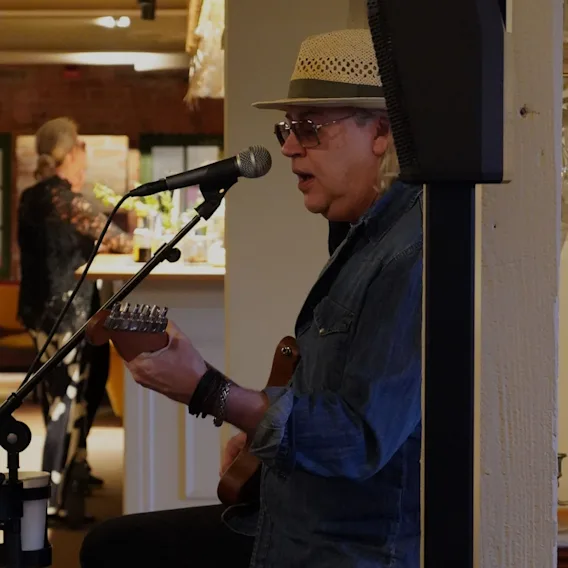 En musiker i stråhatt och solglasögon sjunger i en mikrofon samtidigt som han spelar gitarr.