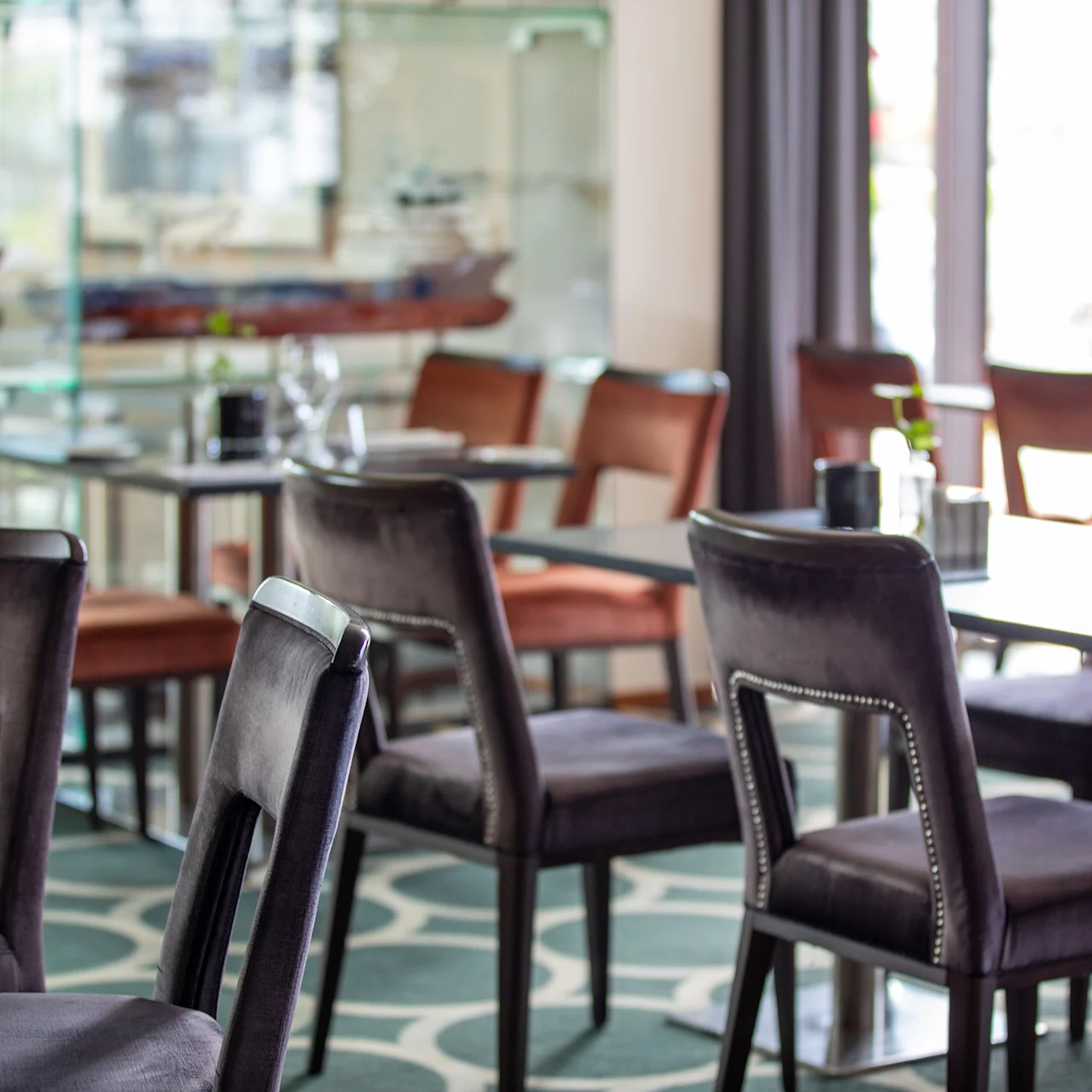 Restaurantstoler omgir bord med glass og bestikk; skip er synlige i et glasskap. Sollys stråler gjennom store vinduer og lyser opp det mønstrede teppet.