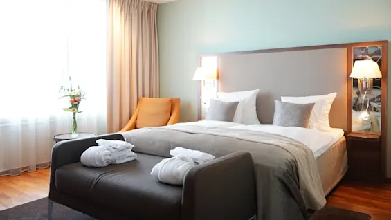 A bed with white and gray pillows stands neatly arranged. Nearby, an orange chair and round table with flowers complete the serene, softly lit room.