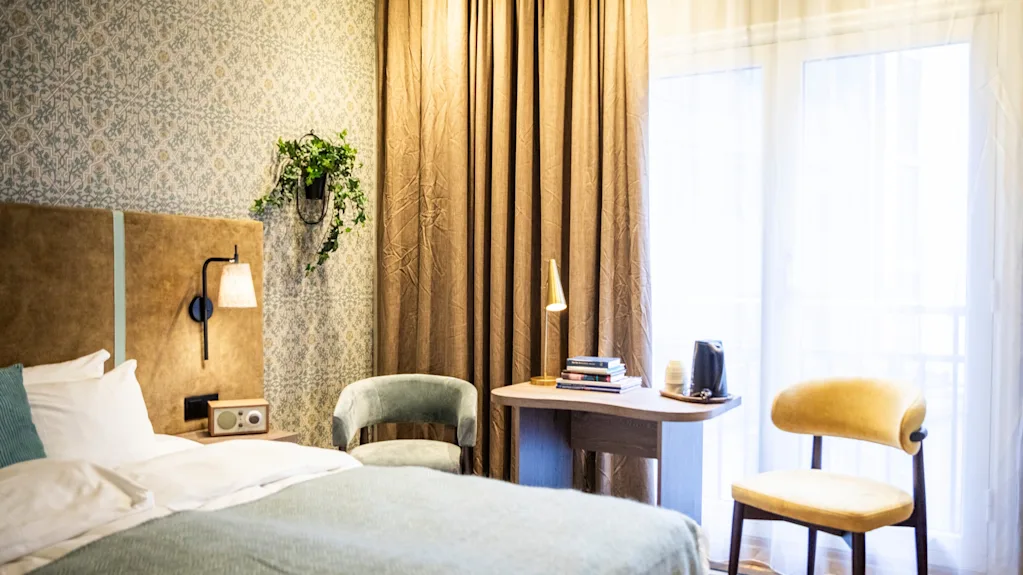 Säng med blå överkast bredvid ett nattduksbord med lampa, i rum med mönstrad tapet, stol, skrivbord och golvlånga gardiner nära ett fönster.