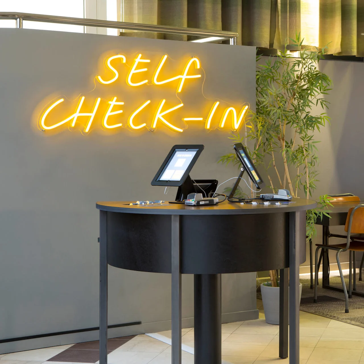 En selvbetjening check-in kiosk med tablets i en moderne lobby, neon "SELF CHECK-IN" skilt ovenfor. Ingen personer til stede.