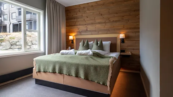 Ett mysigt hotellrum med en säng, trävägg och fönsterutsikt över en byggnad och stenar. Vita handdukar på sängen.