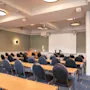 Et konferencelokale med rækker af borde og stole vendt mod en projektorskærm og et podium, klar til en præsentation.