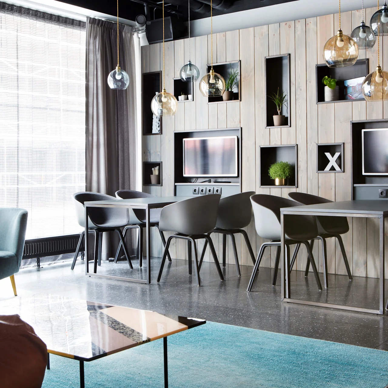 Dining tables with black chairs sit in a modern room, featuring wall shelving, plants, hanging light fixtures, and two televisions against a wood-paneled wall. Large windows illuminate the space.