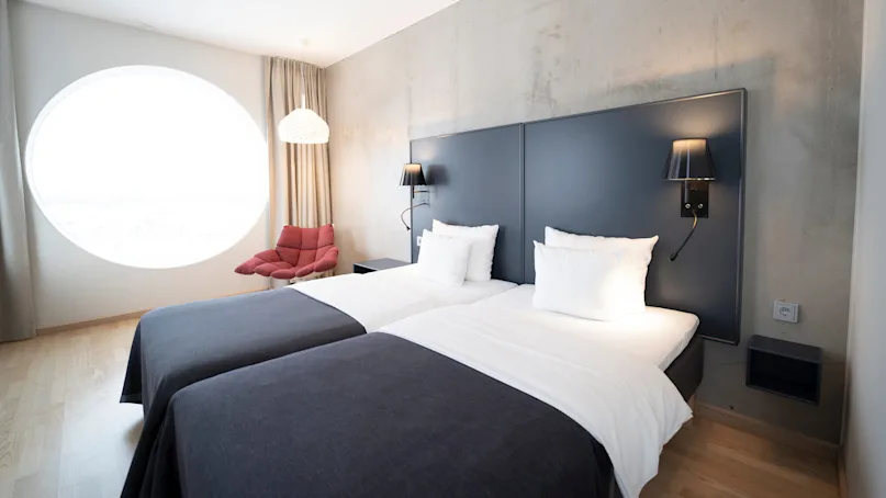 Twin beds with white linens stand against a gray wall, accompanied by black wall lamps. A red chair sits near a large circular window, warmly lit by a hanging lamp.