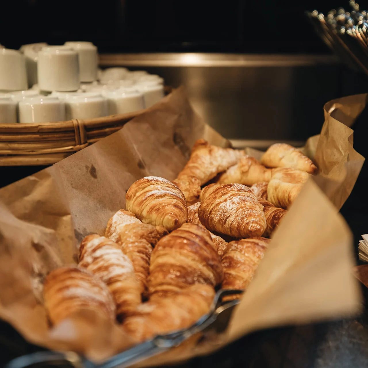 Croissants lie in a paper-lined basket, with white mugs stacked in the background, in a warm bakery atmosphere.