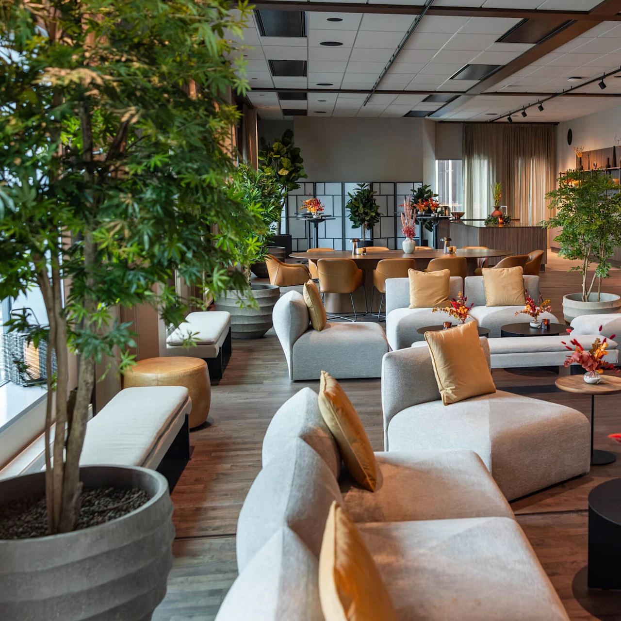 Bekväma sittplatser med beige soffor och gula kuddar är placerade runt svarta soffbord med blomsterdekorationer. Höga krukväxter och en kaffebar i ett träpanelat loungeområde.