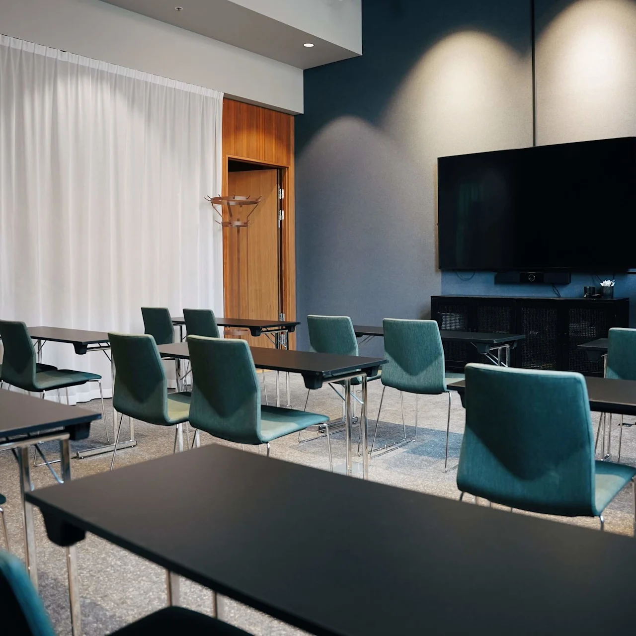 Classroom setup with several rows of black desks and green chairs, facing a large screen and a whiteboard on the wall, surrounded by curtains and wood panels.