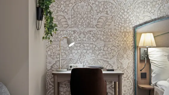 A cozy hotel room featuring a desk with a lamp and chair, a bed with a bedside lamp, and patterned wallpaper.