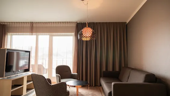 Ett modernt vardagsrum har en grå soffa, två fåtöljer och en TV. Ett runt soffbord står under en hängande pendellampa, med stora fönster och tunga gardiner i bakgrunden.