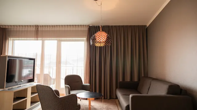 En moderne stue har en grå sofa, to lenestoler og en TV. Et rundt kaffebord står under en hengende pendellampe, med store vinduer og tunge gardiner i bakgrunnen.