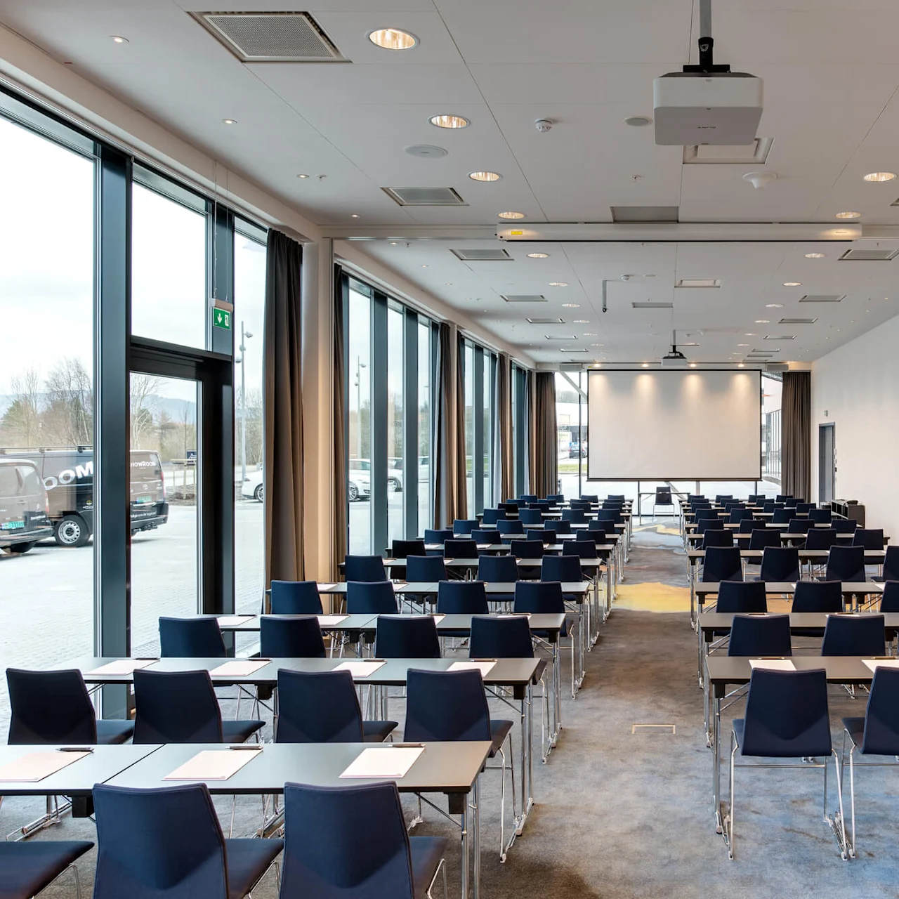 Rows of empty chairs face a large projector screen in a spacious conference room with glass walls. Sheets of paper are placed on the tables, and a digital clock reads 8:02.