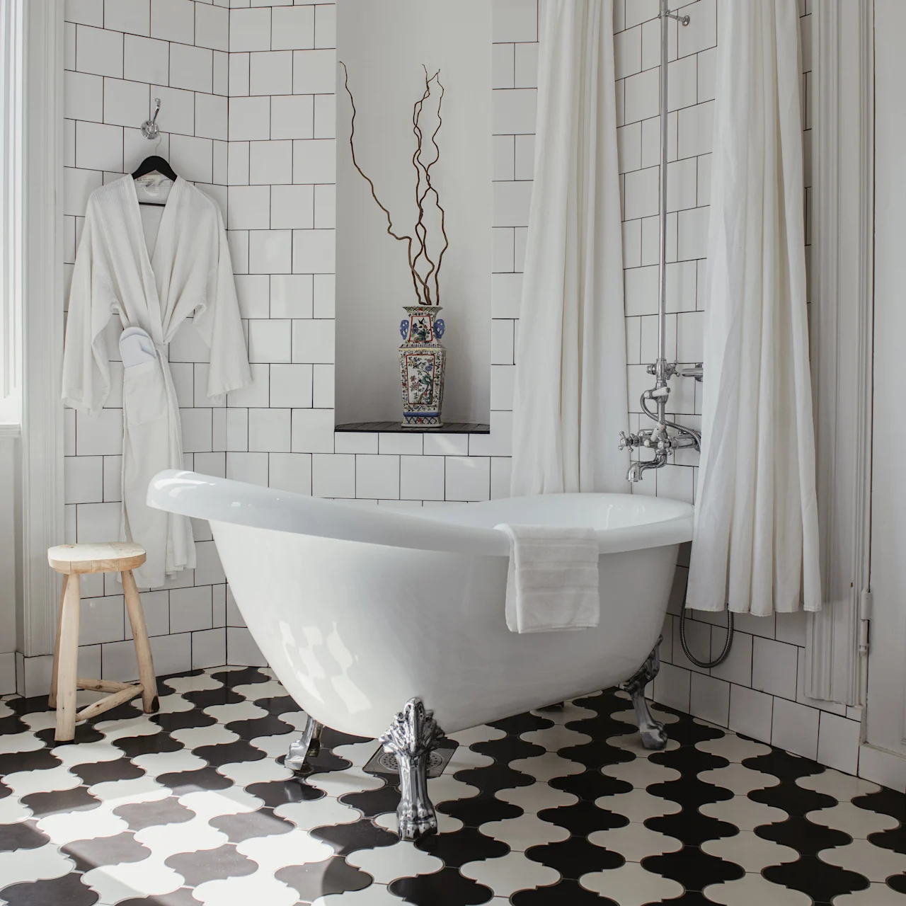 Klofotbadkar står med en handduk draperad över kanten; vit morgonrock hänger i närheten. Svartvitt schackbrädsgolv med kaklade väggar omger det. Radiator och pall ger detaljer.