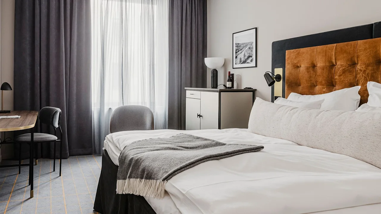 A neatly-made bed with a gray blanket rests in a modern bedroom. Soft light filters through sheer curtains, next to a small desk and chair, enhancing the minimalist decor.