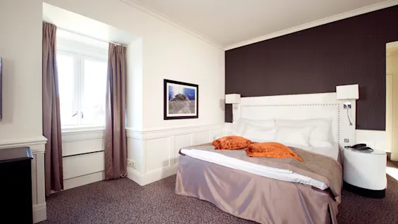 En dubbelsäng med vita kuddar och orange kuddar står mot en mörk fondvägg, flankerad av sängbord, i ett hotellrum med ett fönster och inramad väggkonst.