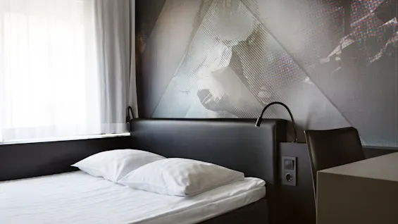 Et moderne hotelværelse med en pænt redt seng med hvide lagner og puder, placeret ved siden af et vindue med tynde gardiner og belyst af vægmonterede læselamper.