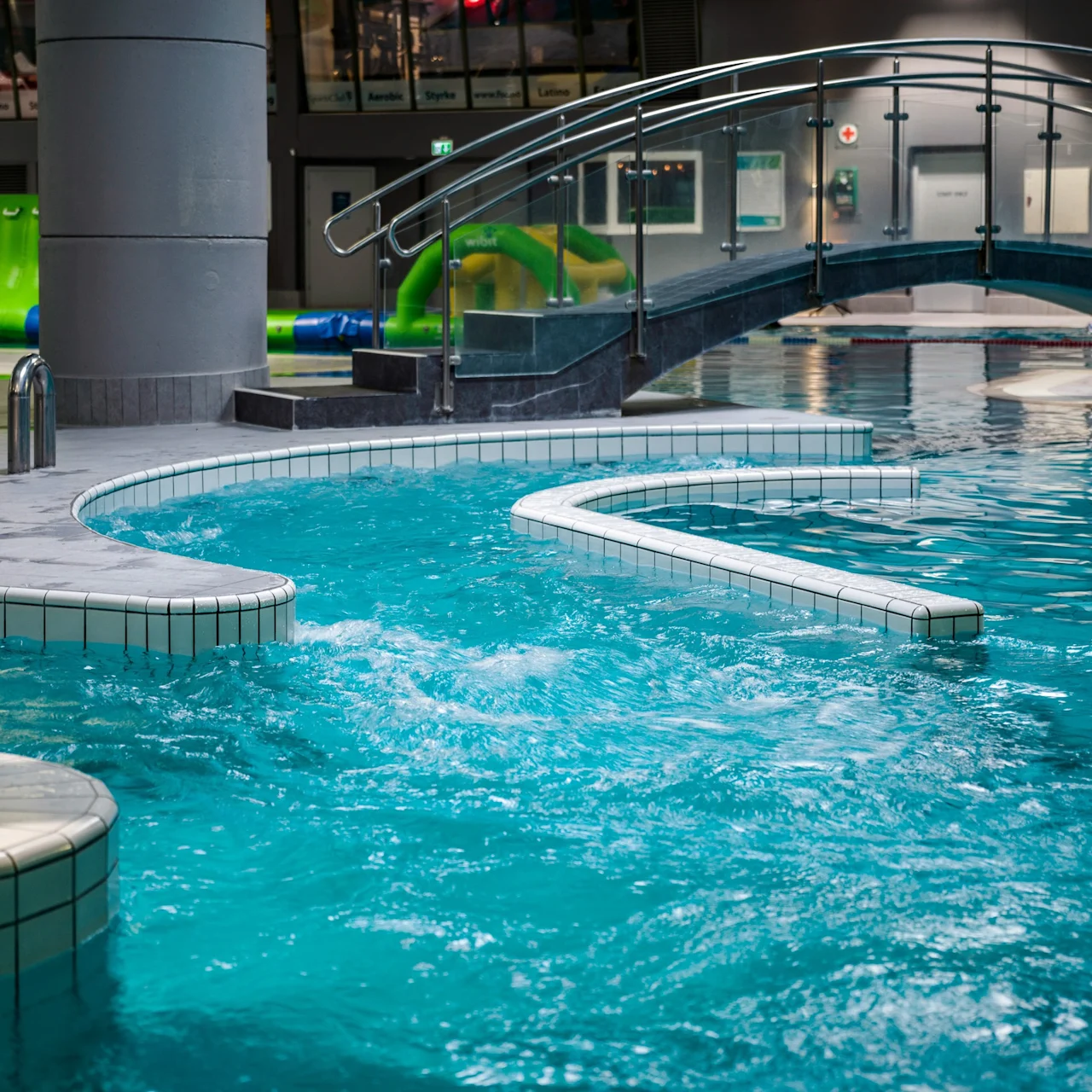 A curved swimming pool with bubbling water is beneath a small arched bridge. In the background, a large green inflatable obstacle course is visible.