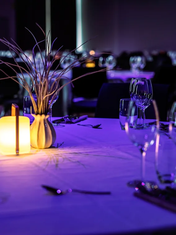 A table set for a formal event features wine glasses and cutlery arranged neatly. A glowing candle and decorative vase with grass create an elegant, ambient atmosphere in a dimly lit room.