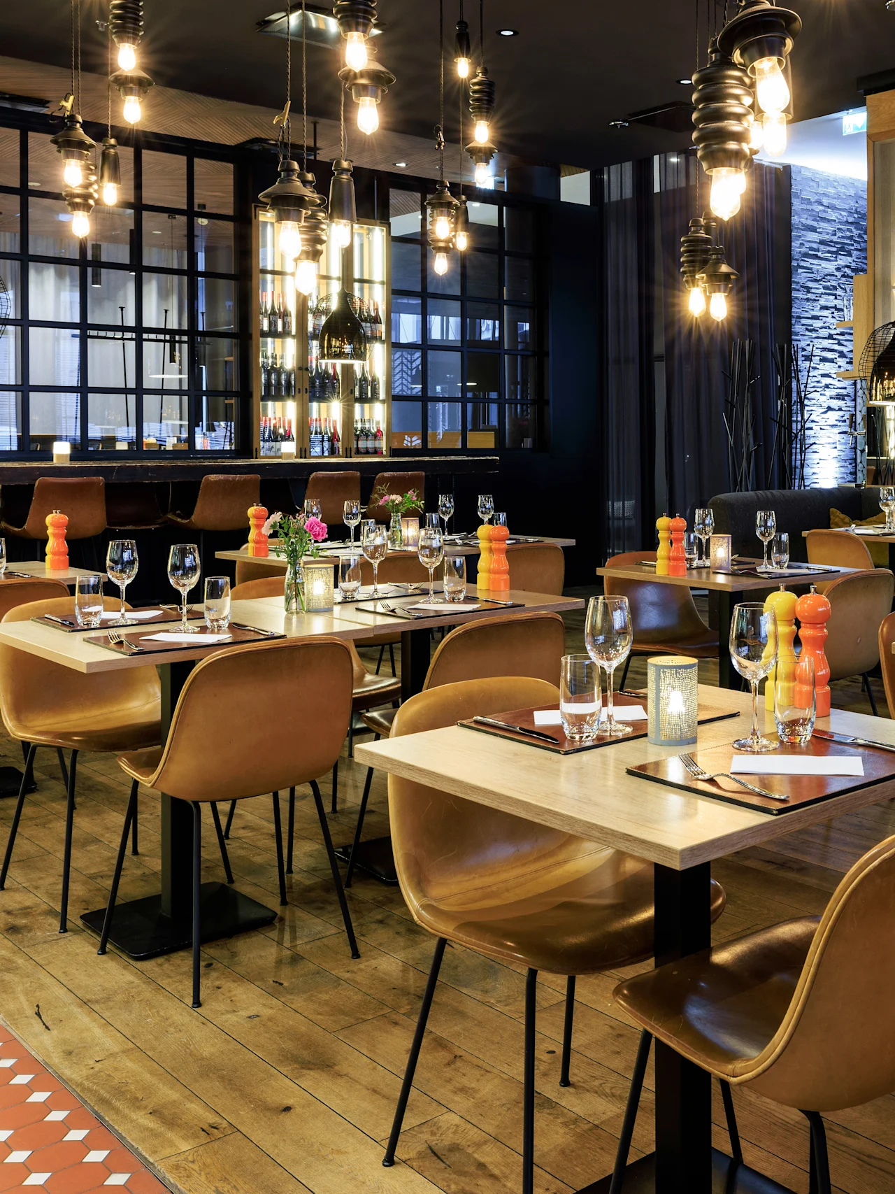 Dining tables set for guests, with leather chairs and elegant glassware, in a warmly lit restaurant. The bar in the background showcases various bottles, adding a cozy ambiance.