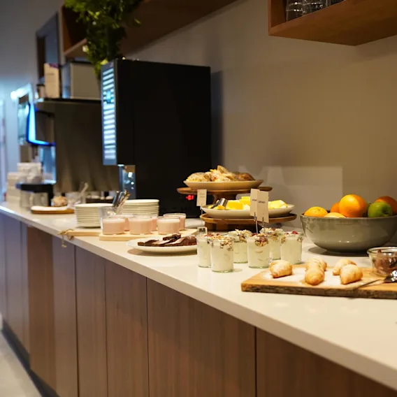 Disken visar olika bakverk, yoghurts, frukter och drycker; placerad bredvid en kaffemaskin och kranar, skapande en frukostbuffé. Ett skylt säger "Laktofri."