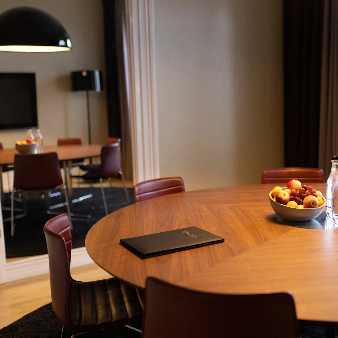 A round wooden table with chairs, a bowl of fruit, and a closed laptop, in a modern meeting room with a view through the window.