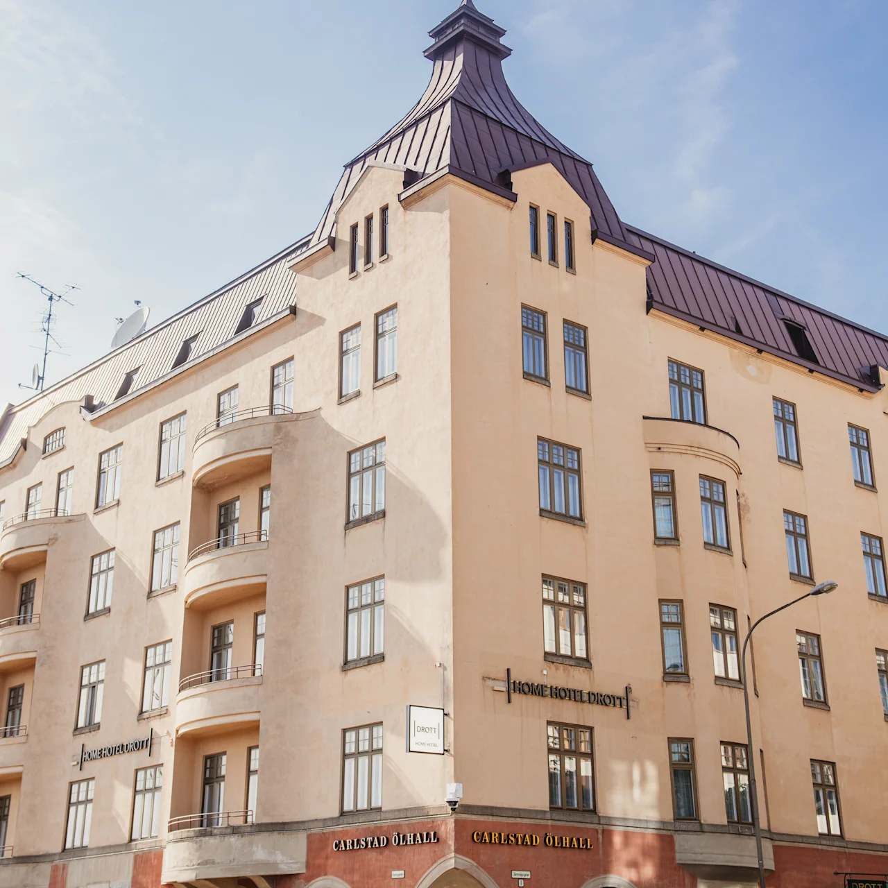 En beige bygning med mørkt tak står på et hjørne. Skilte lyder "HOME HOTEL DROTT" og "CARLSTAD ÖLHALL". Balkoner pryder den ene side.