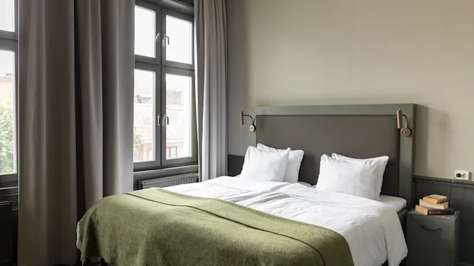 En rolig soveromsscene med en stor, innbydende seng kledd i hvitt og et grønt teppe, ved siden av et vindu med myke grå gardiner.