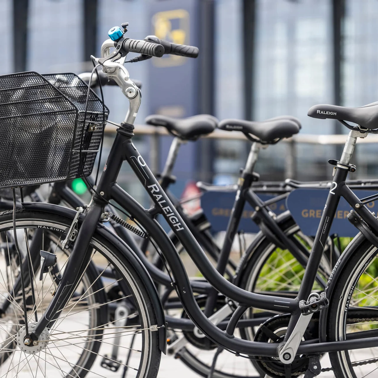 Cyklar med korgar parkerade i rad, märkta "RALEIGH", i ett stadsområde med moderna byggnader i bakgrunden. Text på sidan: "RIDING IN A GREENER."