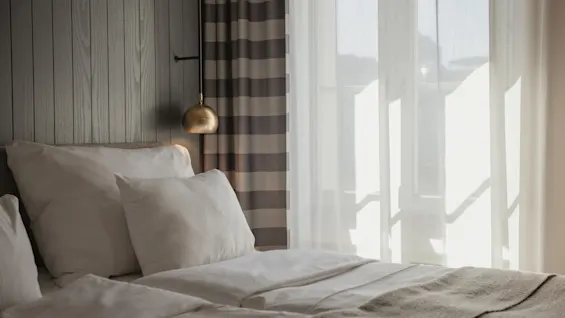 A cozy bedroom with a bed, wooden headboard, and a brass lamp. Sunlight streams through sheer curtains, creating a warm and inviting atmosphere.