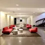 Red sofas sit on a brown rug, surrounded by white stools in a modern lobby with a spiral staircase and overhead lighting.