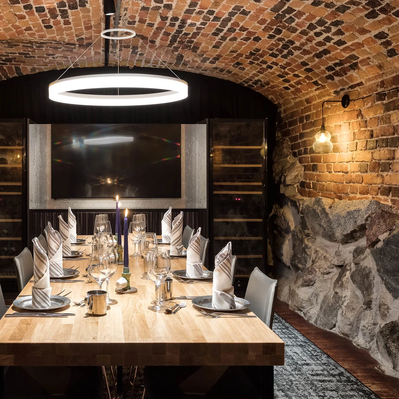 A dining table set for a formal meal in a room with brick and stone walls, ambient lighting, and wine storage.