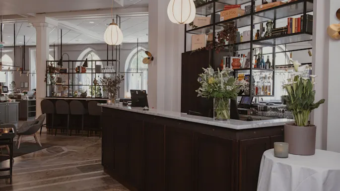 En bardisk pyntet med blomsteroppsatser, i et elegant restaurantinteriør med moderne hengelamper, høye hyller med bøker og flasker, og stilige sitteplasser.
